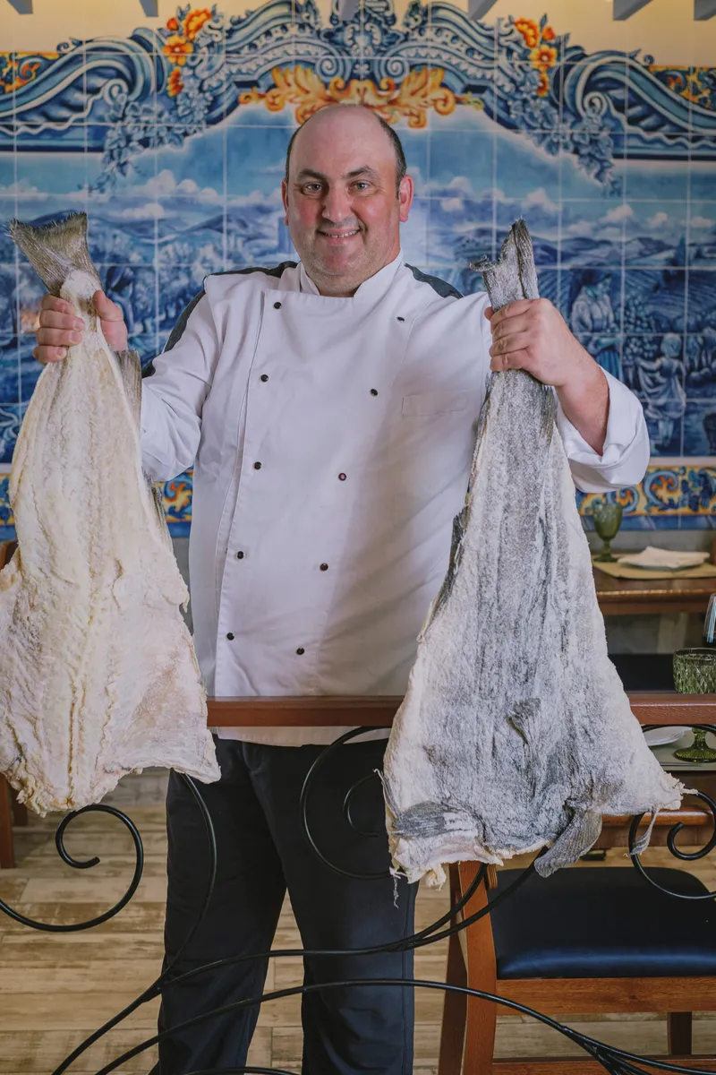 O chef Anderson Antônio segurando dois bacalhaus, diante do painel de azulejos do salão